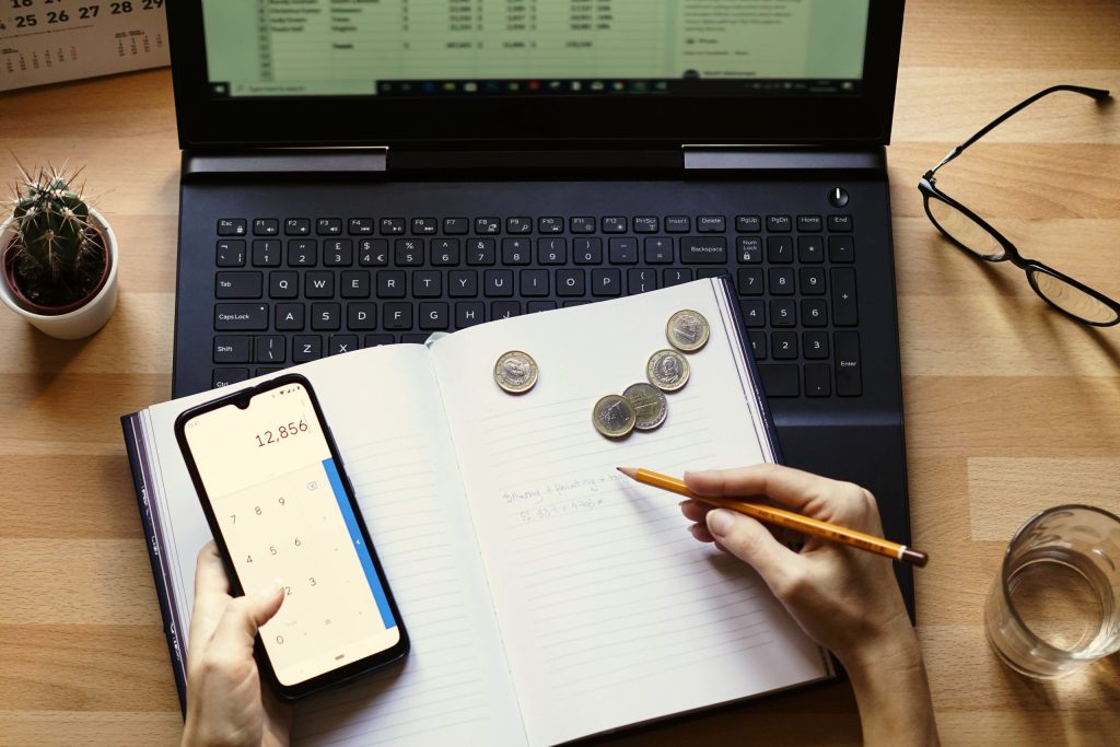 Pessoa calculando valores financeiros com celular, caderno e notebook sobre a mesa