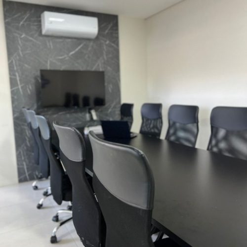 Sala de reunião com mesa longa preta, cadeiras ergonômicas e tela na parede para apresentações