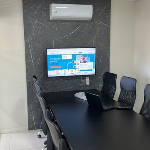 Sala de reunião corporativa com mesa longa, cadeiras ergonômicas, notebook e tela de apresentação na parede